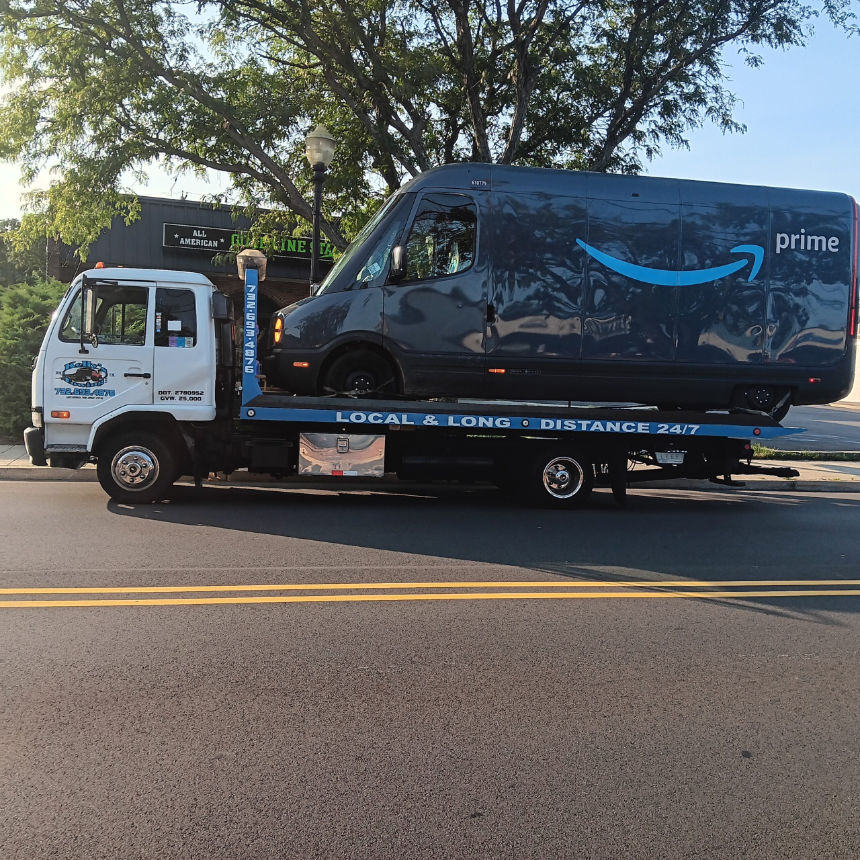 A tow truck transports a blue Amazon Prime delivery van on its flatbed, parked on a street under a tree.