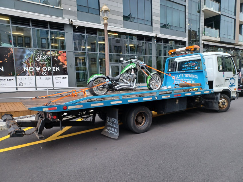 A blue truck carrying a motorcycle in the truck bed, parked on a paved surface.