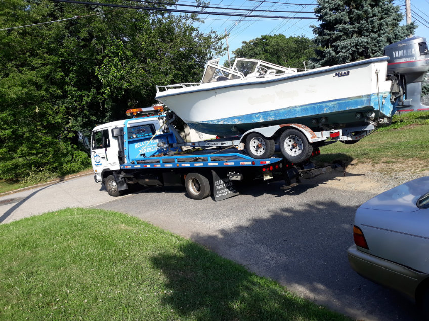 A tow truck transporting a boat on its flatbed, parked on a roadside with trees in the background.