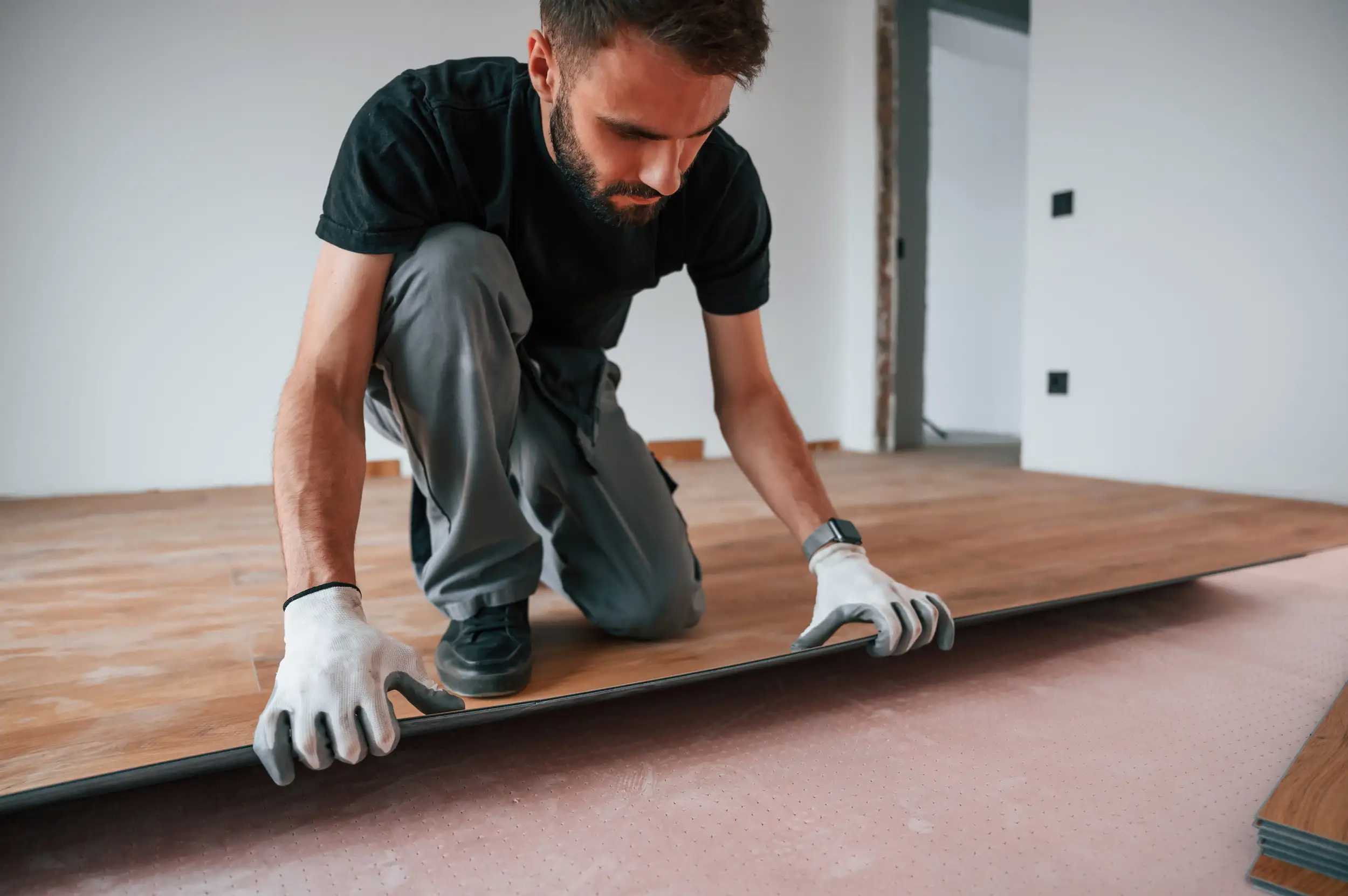 A person wearing gloves kneels to install flooring planks on a wooden surface in a newly renovated room.