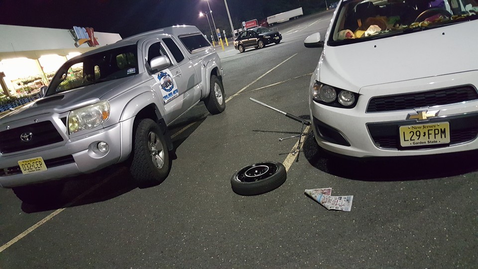 A silver Toyota truck and a white Chevrolet parked at night; the Chevy has a flat tire and a jack, with a newspaper on the ground.