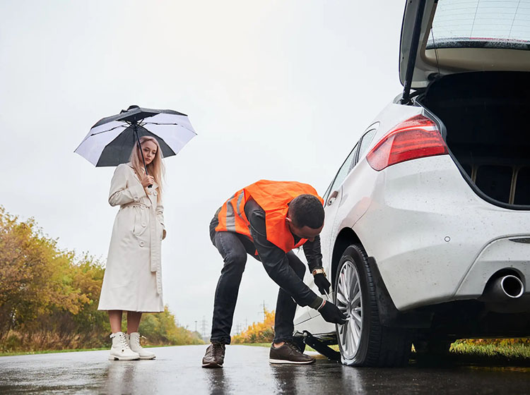 A roadside assistance worker in an orange vest replaces a tire on a white car as a woman in a beige coat holds an umbrella.