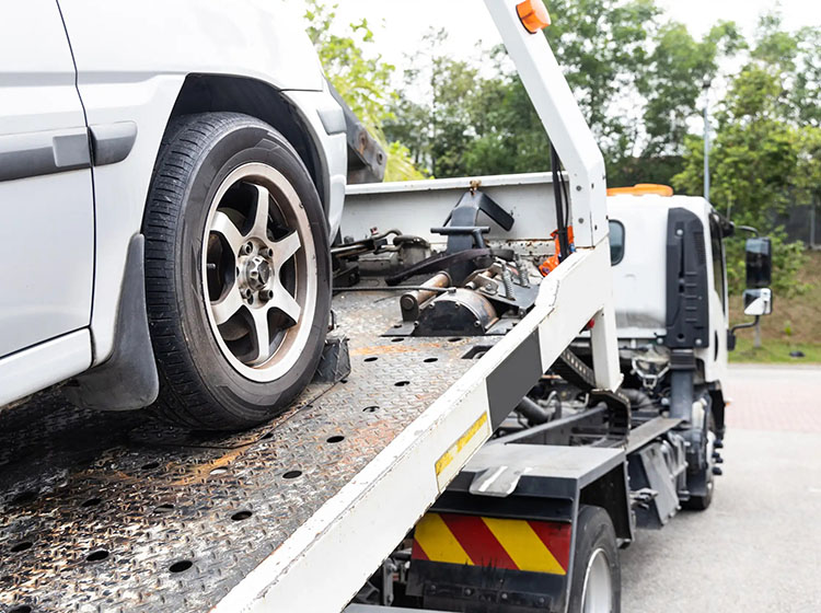 A close-up of a silver car being loaded onto a tow truck, showing its wheel and parts on the flatbed with greenery in the background.